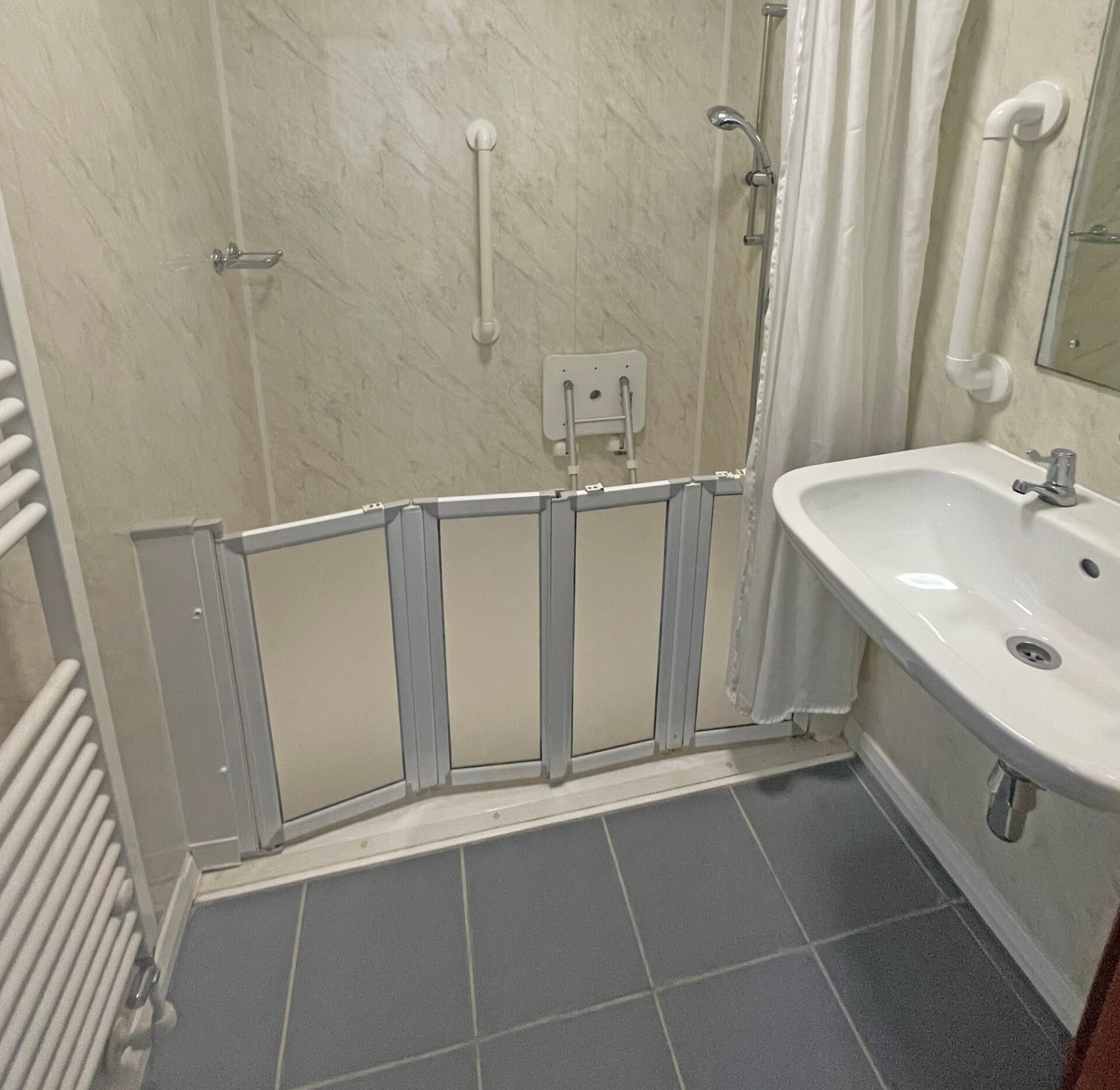 A spacious wet room with white and grey tiled walls, a non-slip floor, and a walk-in shower area. The shower has a handheld and overhead fixture, with a grab rail mounted on the wall. A white fold-down shower seat is attached to the wall for accessibility. To the right, there is a white sink with a mirror above it, and a towel rail mounted nearby. The room is well-lit and designed for accessibility and ease of use.